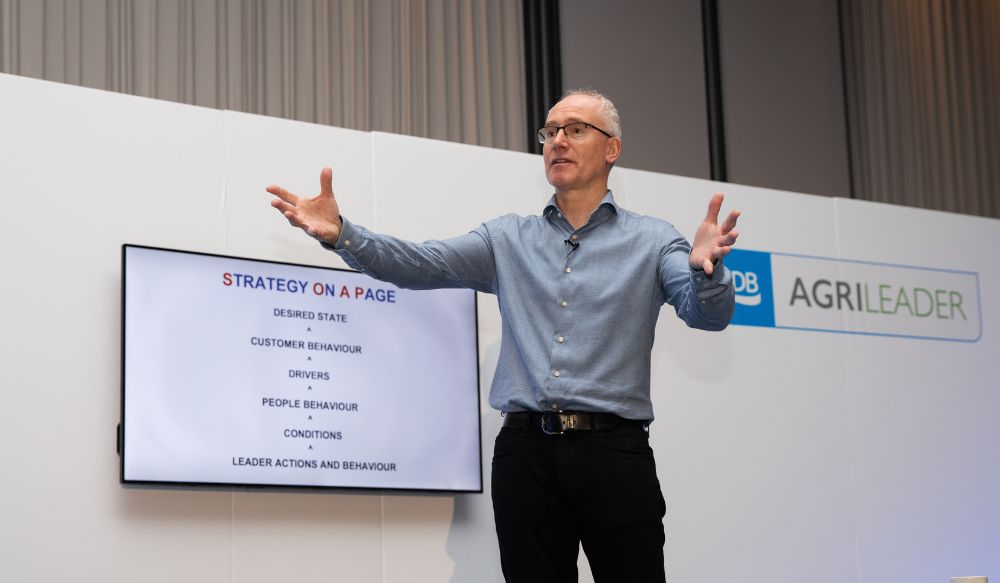 A man (Marcus Child) wearing a blue shirt and dark trousers presenting on stage